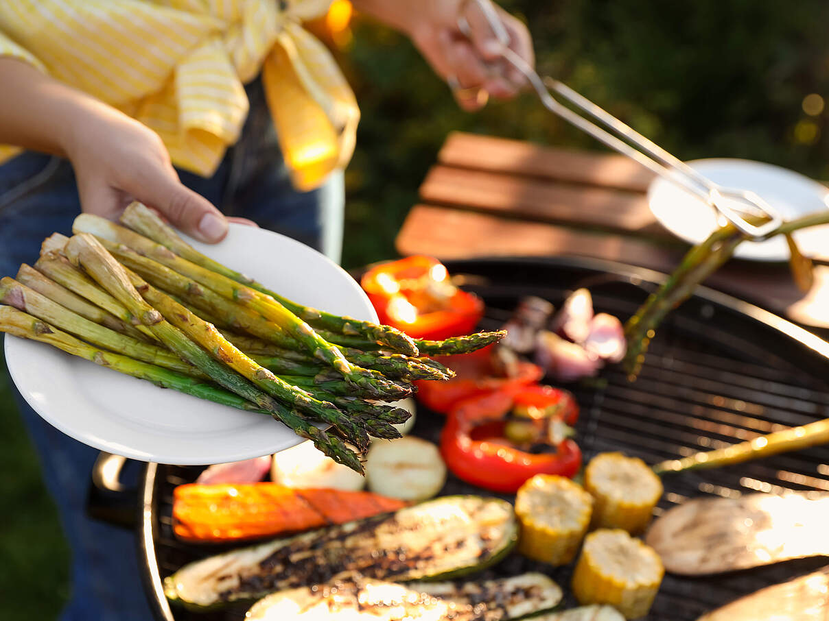 Nahaufnahme einer Person die Gemüse auf dem Grill draussen zubereitet mit einem Teller gegrilltem Spargel im Vordergrund