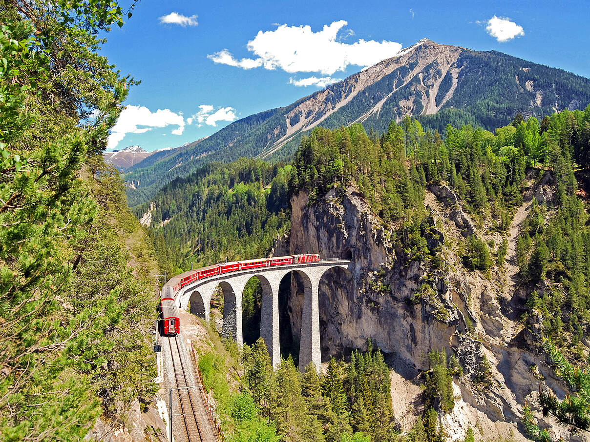 Bahn, die sich auf einem Viadukt durch eine grüne Berglandschaft schlängelt 