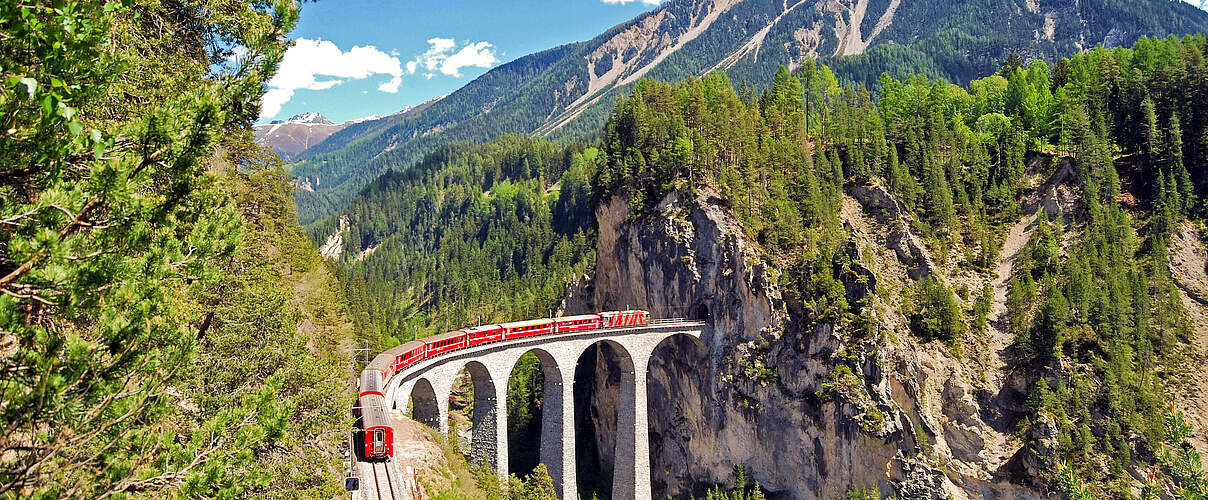 Bahn, die sich auf einem Viadukt durch eine grüne Berglandschaft schlängelt 