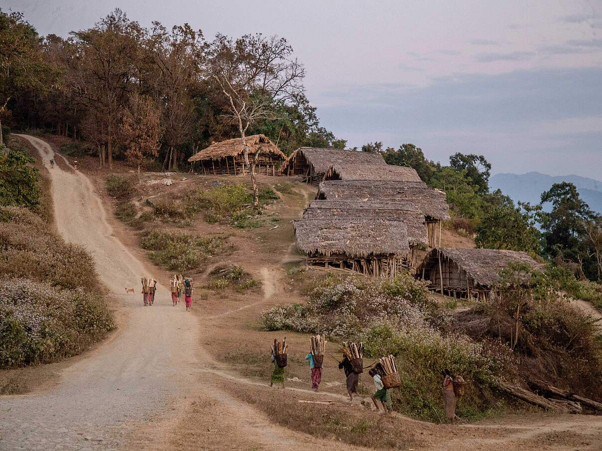 Holzhäuser an Hang, Menschen erklimmen steilen Pfad, auf dem Rücken Tragekörbe mit Holz