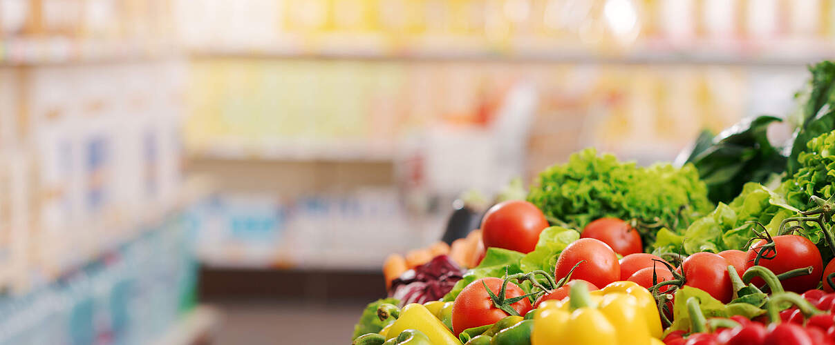 Symbolbild eines Supermarktes mit frischem Gemüse im Vordergrund und einem verschwommenen Hintergrund