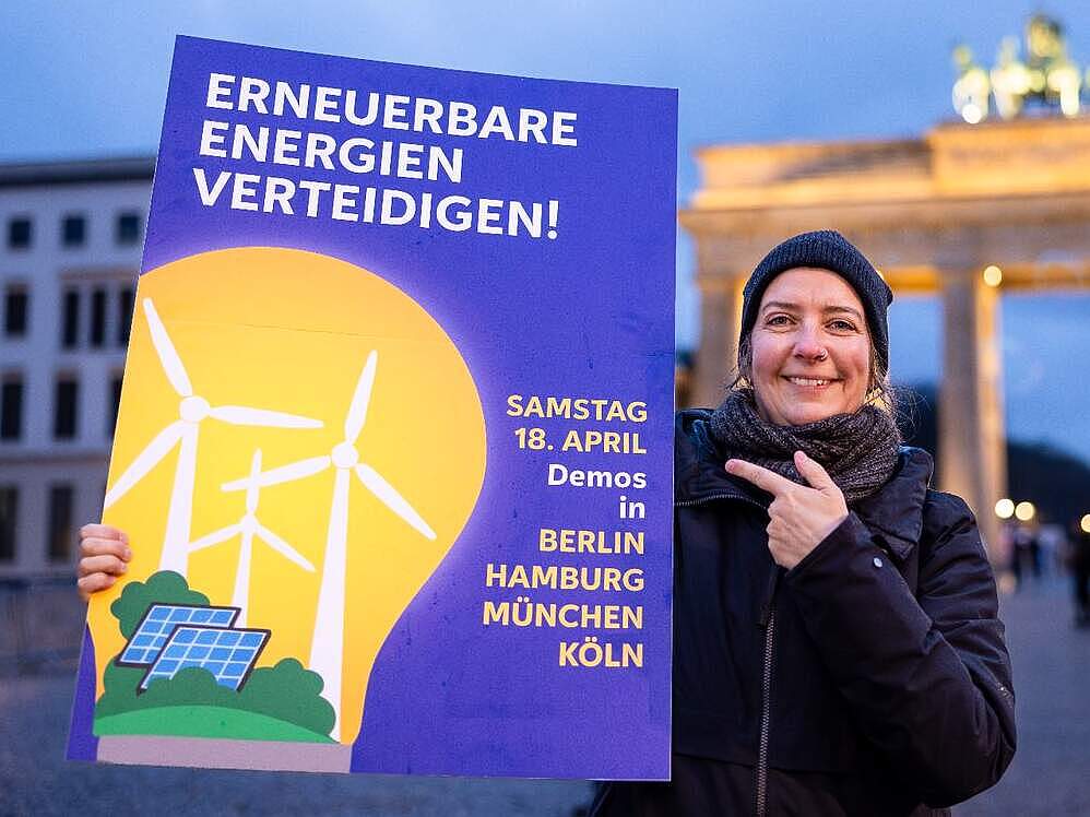 Eine Frau steht vor dem Brandenburger Tor und zeigt auf ein Schild, auf dem „Erneuerbare Energien verteidigen” steht.