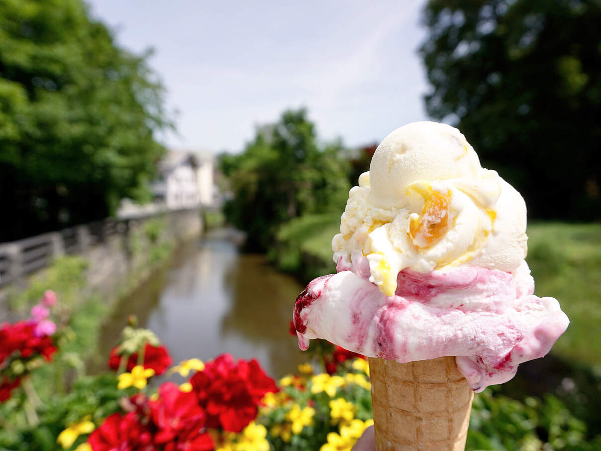 Helles rosa-gelbes Eis in der Waffel wird in die Luft gehalten, drum herum sind leuchtende Frühlingsblumen