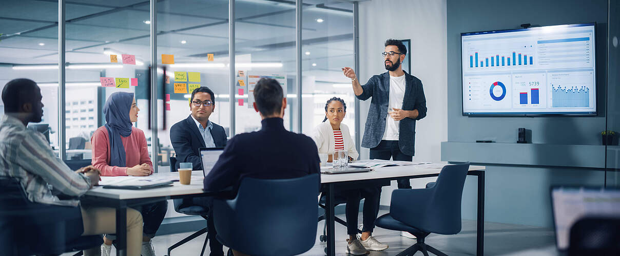 Diverse Modern Office: Businessman Leads Business Meeting with Managers, Talks, uses Presentation TV with Statistics, Infographics. Digital Entrepreneurs Work on e-Commerce Project.