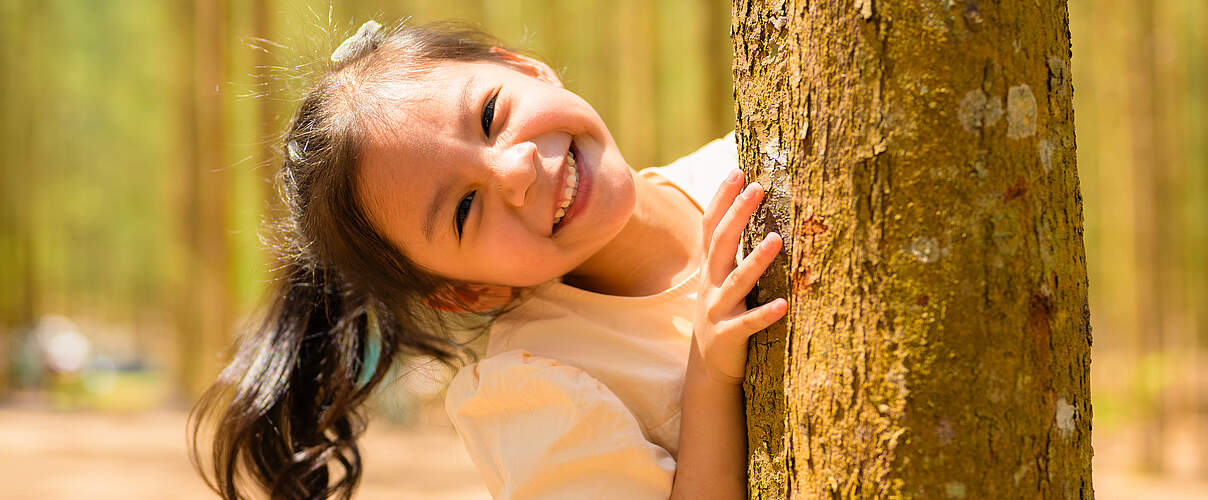Ein Mädchen steht lachend hinter einem Baum 