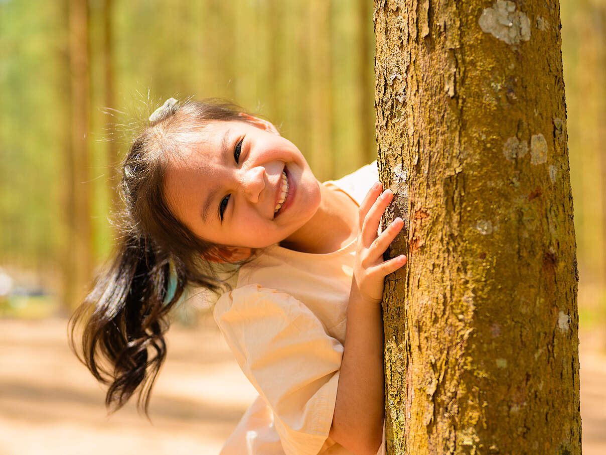 Ein Mädchen steht lachend hinter einem Baum 